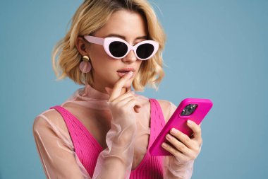 Mavi duvarda izole edilmiş cep telefonu kullanan güneş gözlüklü, kafası karışmış genç bir kadın.