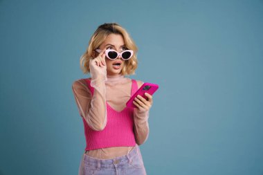 Mavi duvardan izole edilmiş cep telefonu kullanan genç beyaz kadın şaşkınlığını dile getiriyor.