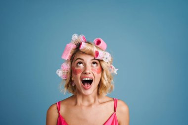 Mavi duvardan izole edilmiş, göz bantları ve bigudileri olan genç bir kadın.