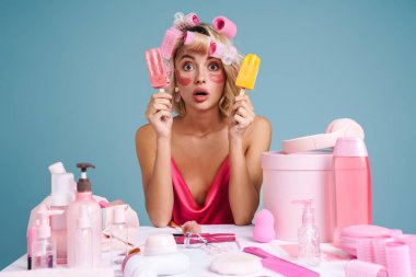Mavi duvarda izole edilmiş güzellik ürünleriyle masaya otururken elinde dondurma tutan sürpriz bir kadın.