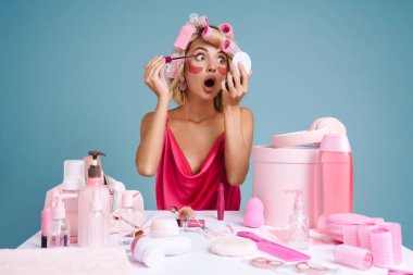 Mavi duvarın üzerinde tek başına otururken bigudili genç bir kadın makyaj yapıyor.