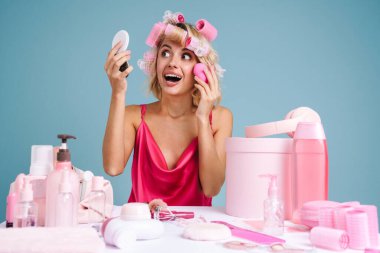 Mavi duvarın üzerinde tek başına otururken bigudili genç bir kadın makyaj yapıyor.