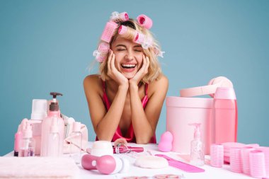 Genç bir kadın, mavi duvarda izole edilmiş güzellik ürünleriyle masada otururken gülüyor.