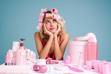 Mavi duvarın üzerinde izole edilmiş güzellik ürünleriyle masaya oturan genç bir kadın yukarı bakıyor.