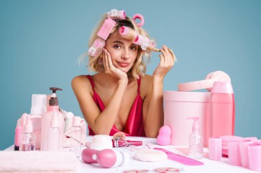 Masada oturmuş gülümseyen genç bir kadın mavi duvarda izole edilmiş güzellik ürünleriyle.