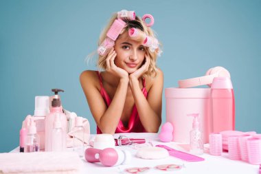 Masada oturmuş gülümseyen genç bir kadın mavi duvarda izole edilmiş güzellik ürünleriyle.