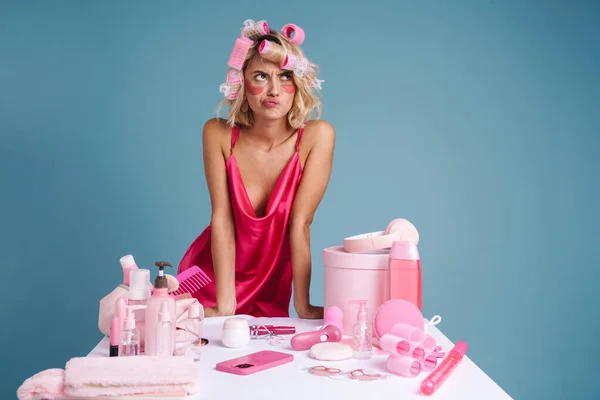 Young woman frowning while standing by table with beauty products isolated over blue wall