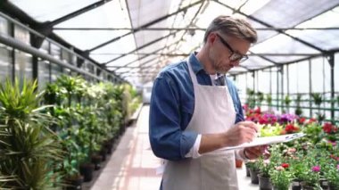 Düşünceli gri saçlı çiçekçi serada kağıda yazı yazıyor.
