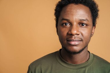 Tişört giyen genç siyah adam bej arka planda izole edilmiş kameraya bakıyor ve poz veriyor.