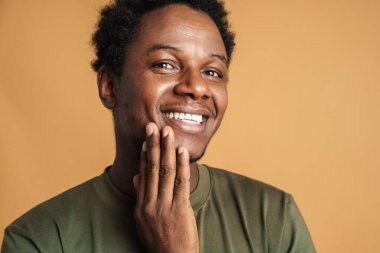 Genç siyah adam tişört giyiyor gülümsüyor ve bej arka planda izole edilmiş kameraya bakıyor.