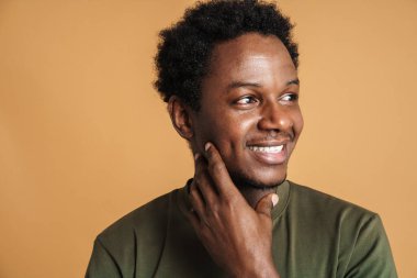 Tişört giyen genç siyah adam gülümsüyor ve bej arka planda izole edilmiş bir yere bakıyor.