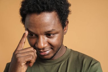Tişört giyen genç siyahi adam bej arka planda poz veriyor ve aşağıya bakıyor.