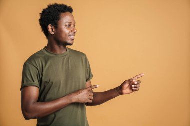 Tişört giyen genç siyah adam gülümsüyor ve bej arka planda izole edilmiş parmakları işaret ediyor.