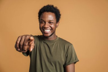 Tişört giyen genç siyah adam gülümsüyor ve bej arka planda izole edilmiş kamerayı işaret ediyor.