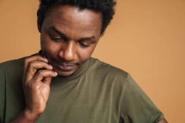 Tişört giyen genç siyahi adam bej arka planda poz veriyor ve aşağıya bakıyor.