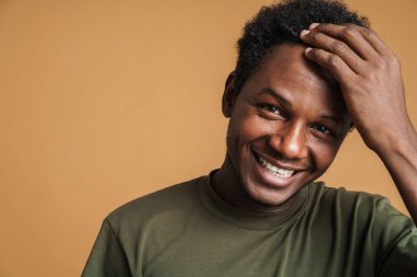 Kafasını tutup bej arka planda izole edilmiş kameraya gülen genç bir zenci.