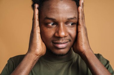 Kafasını tutup bej arka planda izole bir şekilde duran genç siyah bir adam.