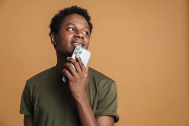 Tişört giyen, gülümseyen ve cep telefonu kullanan genç siyah bir adam bej arka planda izole edilmiş.