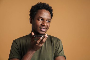 Tişört giyen, gülümseyen ve cep telefonuyla konuşan genç bir zenci bej arka planda izole edilmiş.