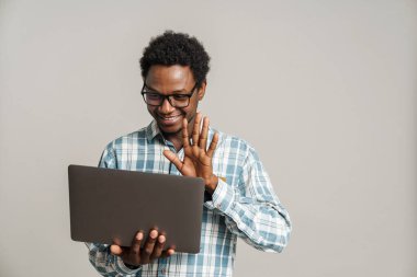 Beyaz arka planda izole edilmiş dizüstü bilgisayar kullanırken gülümseyen ve el kol hareketi yapan genç bir zenci.