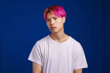 Pembe saçlı Asyalı çocuk somurtuyor ve mavi arka planda izole edilmiş kameraya bakıyor.