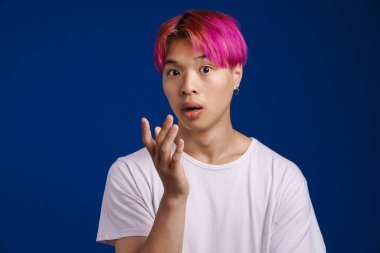 Pembe saçlı Asyalı çocuk mavi arka planda izole edilmiş kameraya şaşırdığını ifade ediyor.