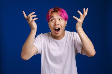 Pembe saçlı Asyalı çocuk mavi arka planda izole edilmiş kameraya şaşırdığını ifade ediyor.