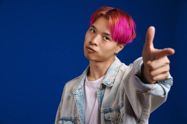 Pembe saçlı Asyalı çocuk mavi arka planda izole edilmiş kameraya işaret ediyor.