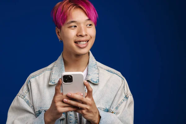 デニムジャケットに身を包んだアジアの少年は 青い背景の上に隔離された携帯電話を使いながら微笑む — ストック写真
