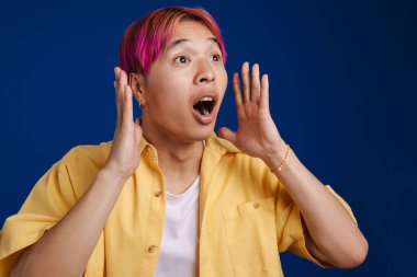 Pembe saçlı Asyalı çocuk mavi arka planda izole edilmiş kameraya şaşırdığını ifade ediyor.