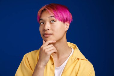 Pembe saçlı Asyalı çocuk mavi arka planda tek başına poz veriyor ve bakıyor.