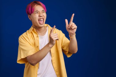 Pembe saçlı, bağıran ve parmaklarını yukarı gösteren Asyalı çocuk mavi arka planda izole edilmiş.