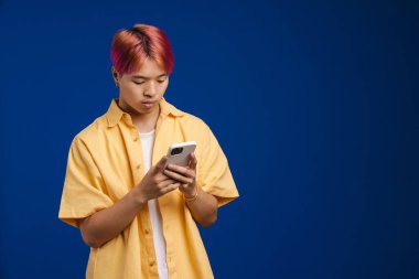 Pembe saçlı Asyalı çocuk mavi arka planda kamerada poz verirken cep telefonu kullanıyor.