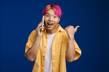 Asyalı çocuk mavi arka planda izole bir şekilde cep telefonuyla konuşurken parmağıyla işaret ediyor.