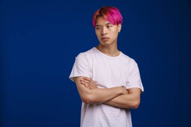 Pembe saçlı Asyalı çocuk mavi arka planda tek başına poz veriyor ve bakıyor.