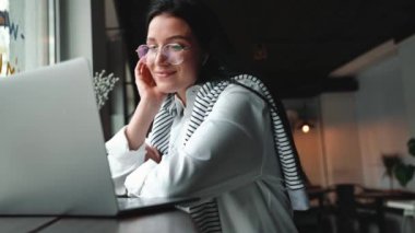 Gözlüklü, gülümseyen esmer kadın kafede bilgisayarla konuşuyor.