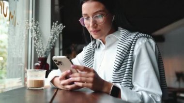 Gözlüklü yakışıklı esmer kadın kafede cep telefonunda yazı yazıyor.