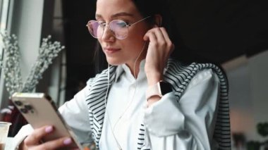 Gözlüklü neşeli esmer kadın kafede cep telefonunda yazı yazıyor.