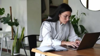 Gözlüklü genç esmer kadın dizüstü bilgisayarda çalışıyor ve kafede not defterine yazıyor.