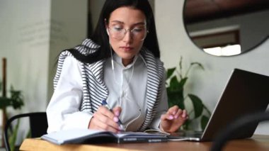 Gözlüklü, laptopta çalışan ve kafede not defterine yazan esmer bir kadın.