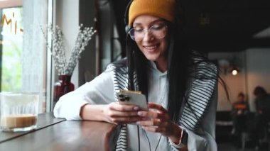 Şapkalı ve gözlüklü güzel esmer kadın kafede telefonla mesajlaşıyor.