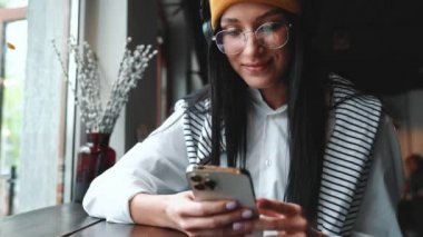 Yakışıklı, esmer, şapkalı, gözlüklü, kafede telefonla mesajlaşan kadın.