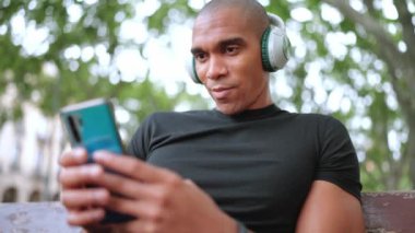 Mutlu kel Afrikalı adam kulaklıkla müzik dinliyor ve dışarıda bankta otururken telefonla yazı yazıyor.