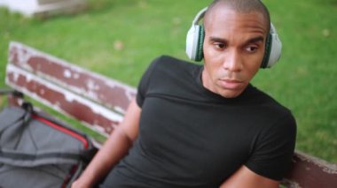 Yakışıklı, kel Afrikalı bir adam bankta otururken kulaklıkla müzik dinliyor.
