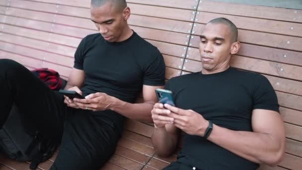 Twee Ontspannen Kale Afrikaanse Broers Typen Telefoons Terwijl Buiten Bank — Stockvideo