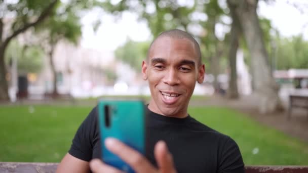 一个快乐的秃头非洲男人坐在外面的长椅上 通过电话视频交谈 并表现出了贝尔利索的手势 — 图库视频影像