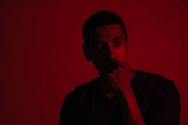 Genç siyahi adam çenesini tutuyor ve kırmızı duvarın üzerinden tek başına bakıyor.