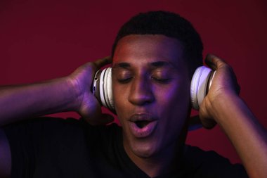 Genç siyahi adam, kırmızı duvarda izole edilmiş kulaklıklarla müzik dinlerken şarkı söylüyor.