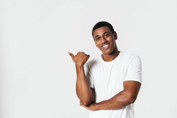 白い壁の上に隔離されたTシャツの笑顔と指差す指を身に着けている若い黒人男性 — ストック写真