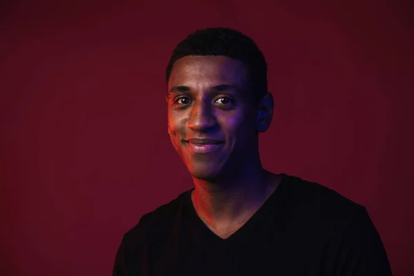 Young black man in t-shirt smiling and looking at camera isolated over red wall